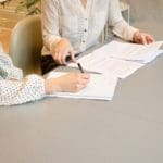 two woman at desk exchange paperwork