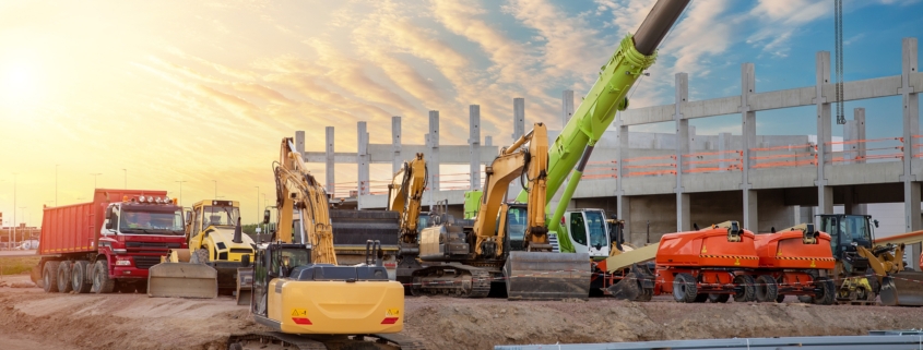 Many different multiclored colorful heavy industrial machinery equipment at construction site parking area against warehouse building city infrastructure development. Commercial vehicles rental sale